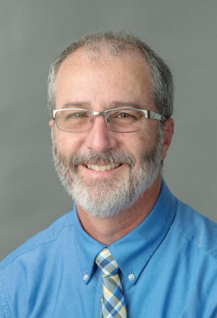 Portrait of a smiling and bearded Christopher Kennedy wearing glasses and a blue shirt and plaid tie.