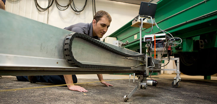 Man on hands and knees examining a piece of equipment that has a long horizontal beam with a belt and other apparatuses attached to it