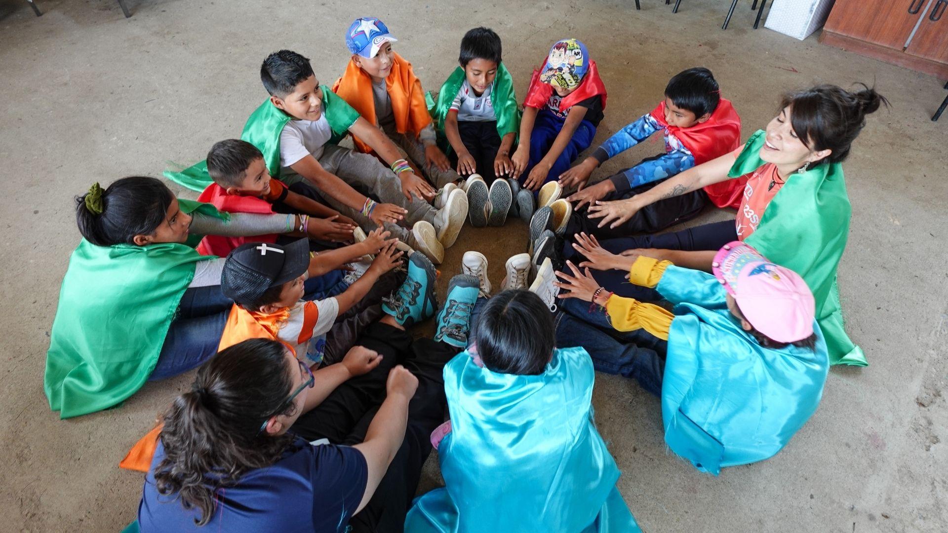 A group of children wearing capes sitting on the floor trying to touch the top of their feet.