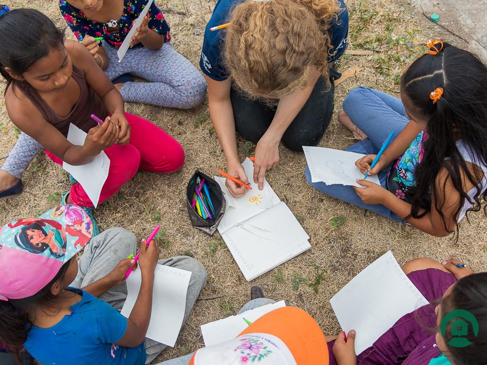 A group of children and a volunteer are drawing on sheets of paper on the ground.