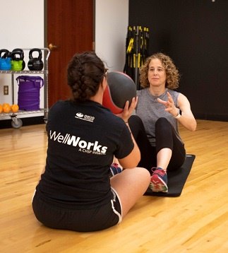 Two members passing a medicine ball between each other in a sitting position with their shoe tips touching and knees against their chest.