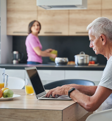 Employee on computer at kitchen table with spouse behind him