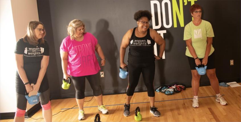 Members smiling and using kettlebells in group fitness room.