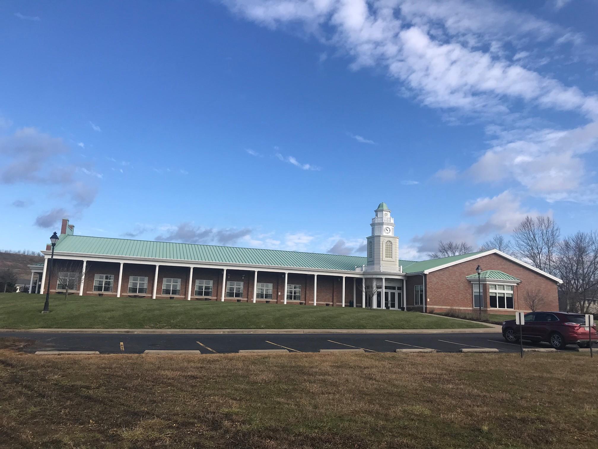 1-story brick building with white columns and clock tower, fronted by a lawn that gently slopes down to an asphalt parking lot