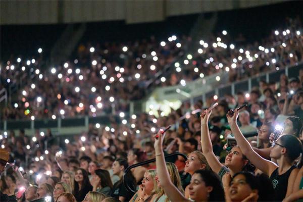 Students hold up cell phones with flashlights turned on in a dark Convocation Center