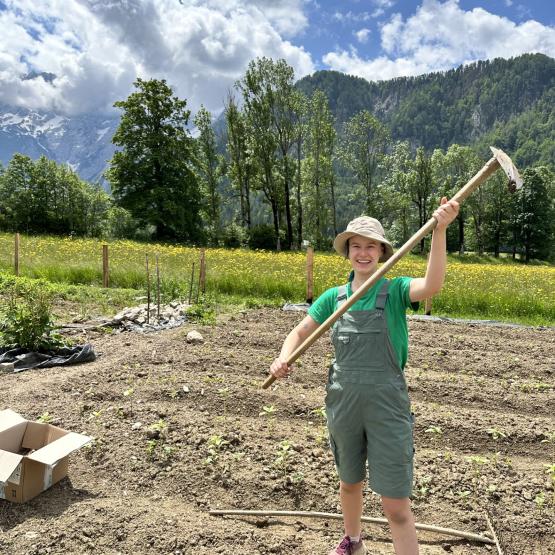 Picture of Celia Hawk in an agricultural field holding a farm tool with mountains and trees in the background.