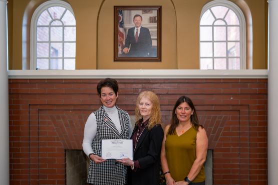 Three women pose for a photo in front of a portrait of George Voinovich. The woman on the left is holding a framed certificate