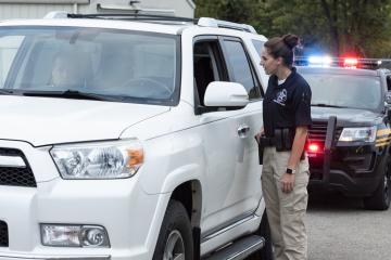 A law enforcement officer is shown standing next to a parked vehicle and speaking with someone in the vehicle.