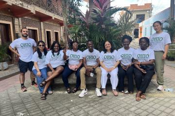 A group of students wearing OHIO t-shirts are seated under a tree, smiling at the camera