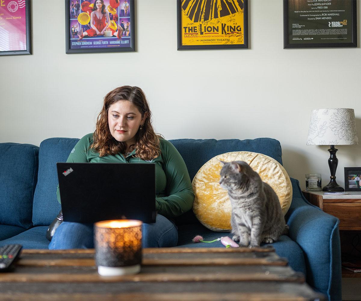 A student works at their laptop while sitting on their couch at home with their cat sitting beside them.