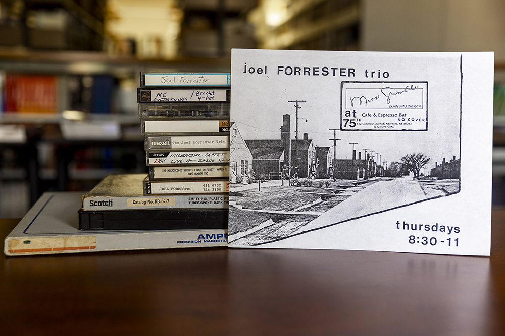 Photo of Forrester's donations of vinyl, tapes, and CDs staked behind a small post card sized advertisement