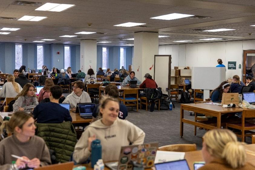 An open floor area filled with wooden tables and chairs densely populated by students