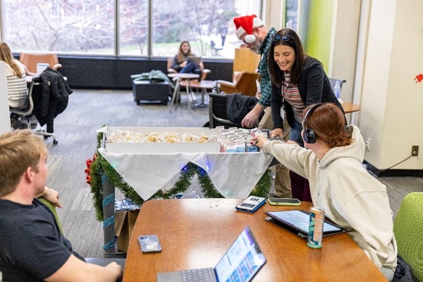 A woman offers milk and cookies from a cart to two students seated at a table