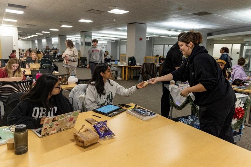 A student employee hands a packaged cookie to a student seated at a table in a crowded study area