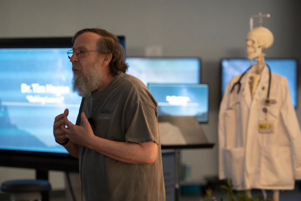 Man speaking with skeleton wearing a white coat in the background