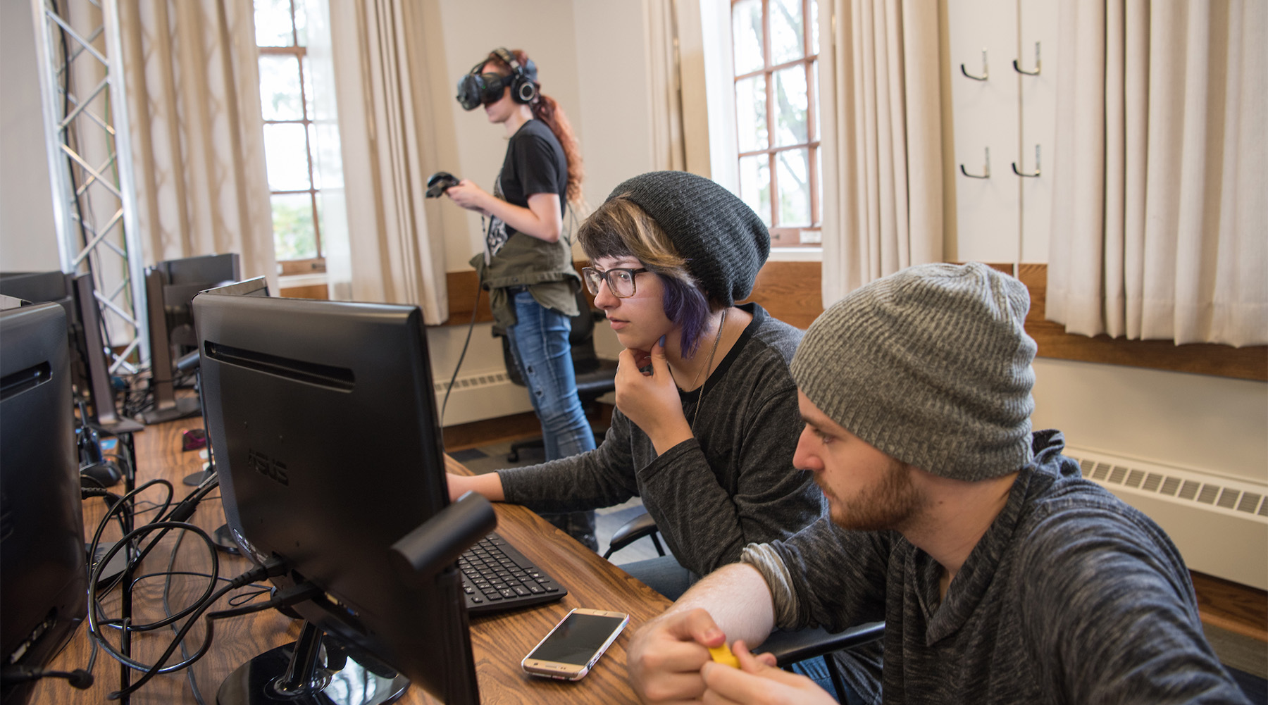 Two students work at a computer while another tests out VR equipment.