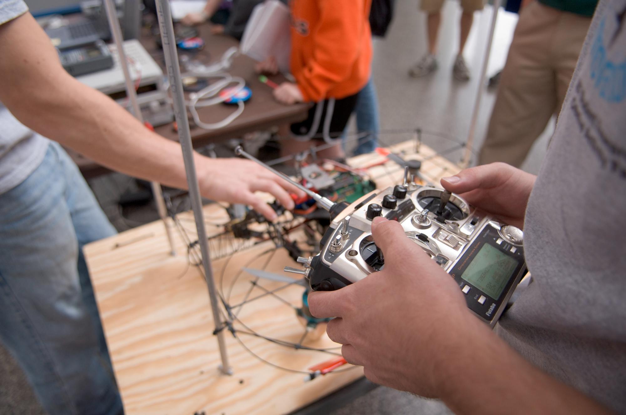 Hands hold a double-joystick remote controller, and while an arm reaches toward wires in the background