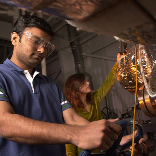 Man grips valve wheel in foreground, woman reaches for duct tube in background