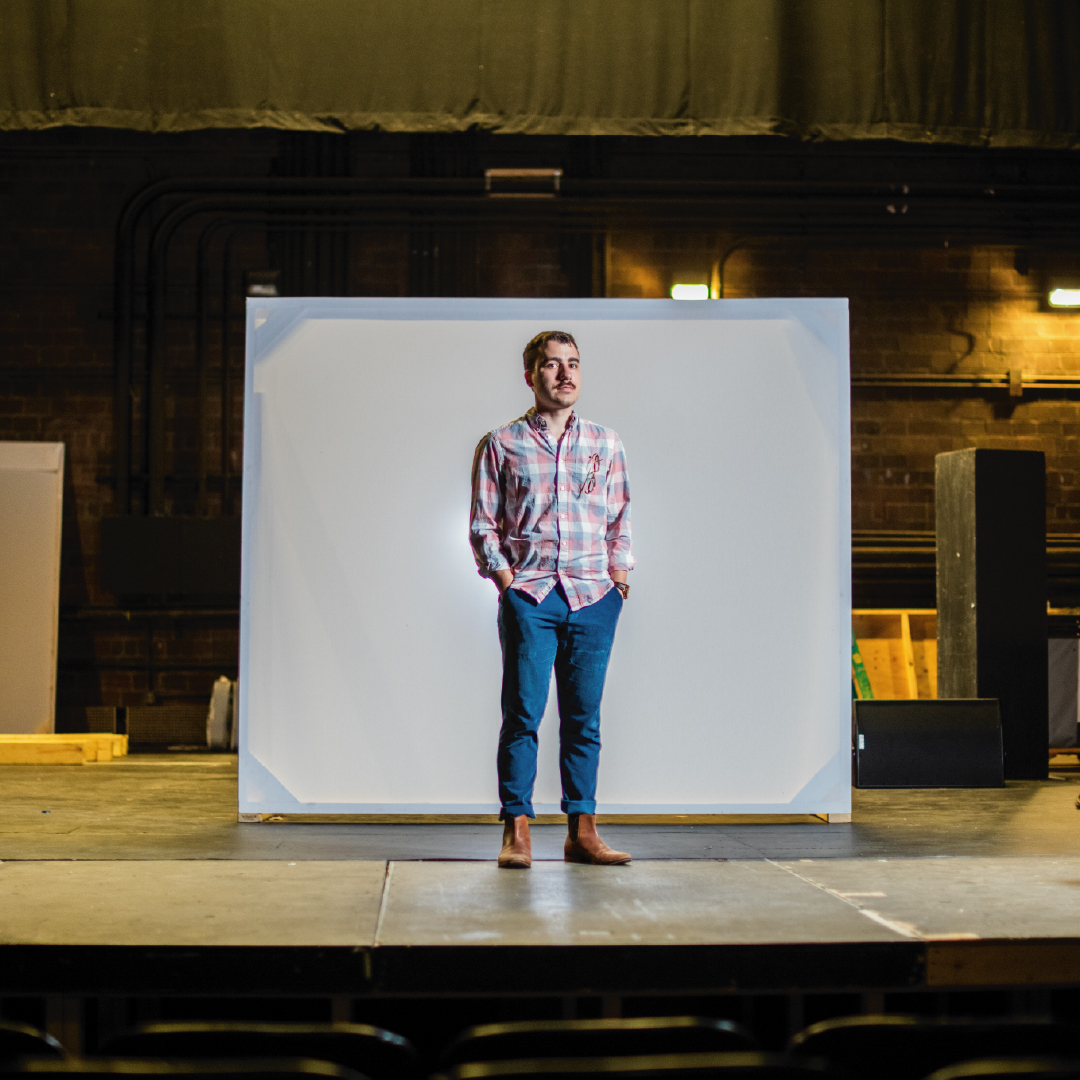 A graduate student poses on a theater stage in front of a backdrop