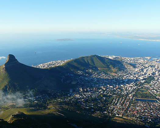 View from mountain looking down on Cape Town. The mountain and water frame the city on the left side of the photo. The water is blue and there is lots of green vegetation surrounding the city.