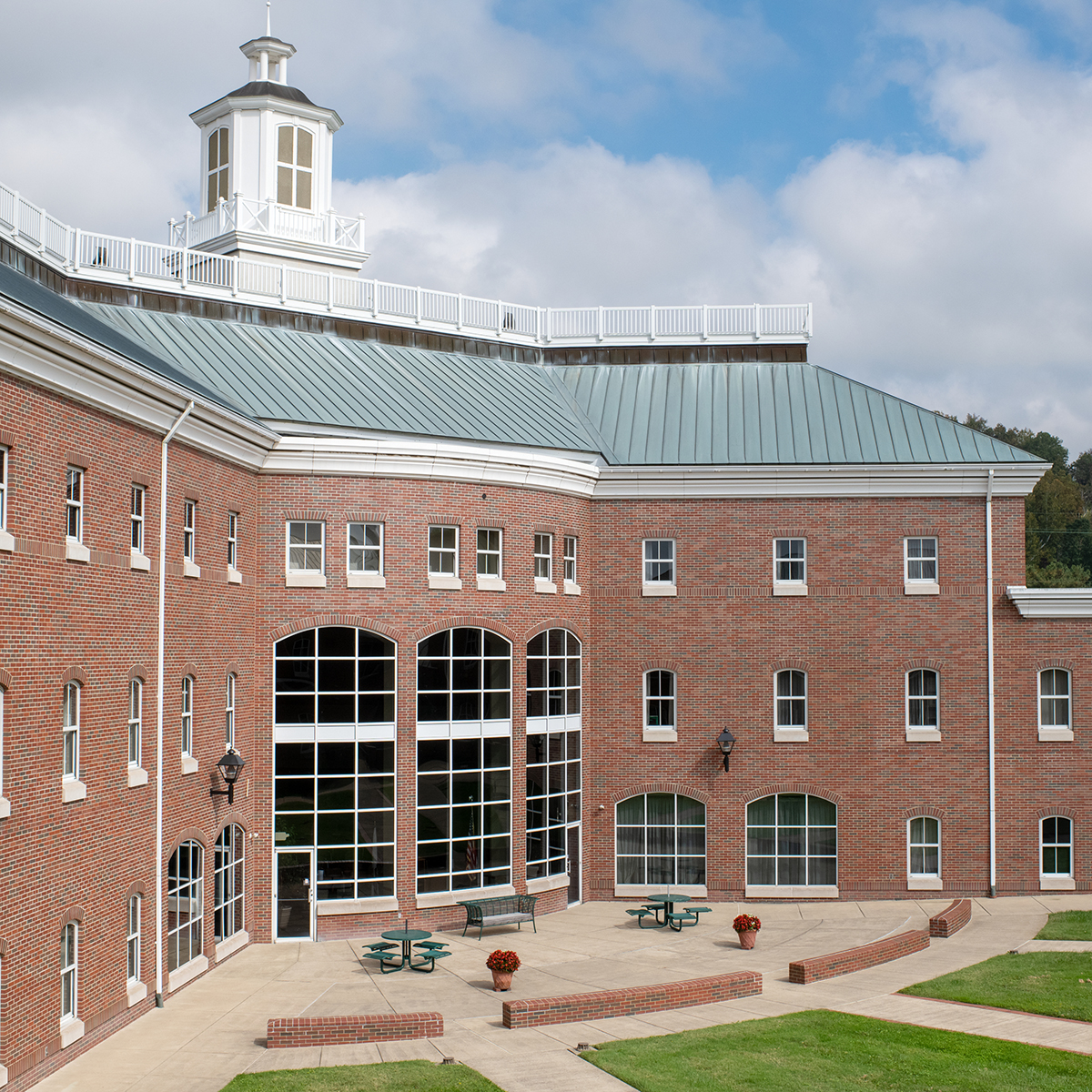 A three-story brick building with many windows. On the middle section, there is a large window that is two stories tall. It has a green tile rood and a white spire.