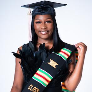 Salih Lewis wears a black gown with a feather-like top, smiling and wearing a sash and a graduation cap
