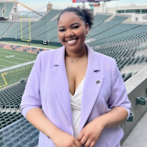 Azaria Greene-Williams stands in front of Peden football field, smiling at the camera and wearing a white tank top and pale purple blazer.
