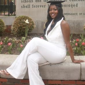 Caila Moniece Gissentaner sits in front of Ohio University's entrance, wearing a white jumpsuit and graduation cap.