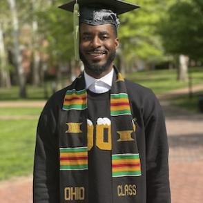 Jalin Fon Holloway stands on college green wearing his graduation cap and gown.
