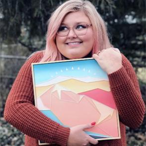 Laura Brooks holds a square piece of art in her hands and smiles, looking away from the camera.