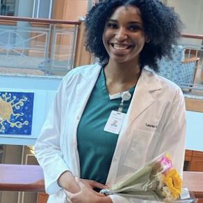Lauren Morine Harris holds a small bouquet of flowers and smiles at the camera, wearing a white coat over her green scrubs.