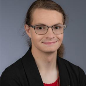 Luvina Cooley half-smiles, standing in front of a grey background and wearing a red shirt and black cardigan with a transgender pride pin on the lapel