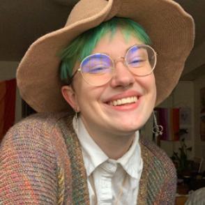 Owen Yates-McEwan smiles at the camera in a wide-brimmed tan hat, white collared shirt, golden rimmed glasses and a multi-colored cardigan