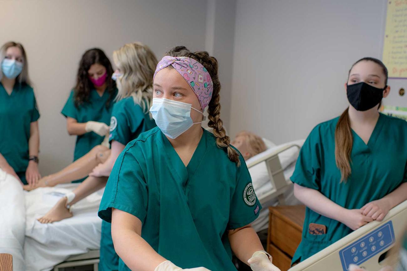 A nursing student near a patient bed looks off-camera while other nursing students stand in the background