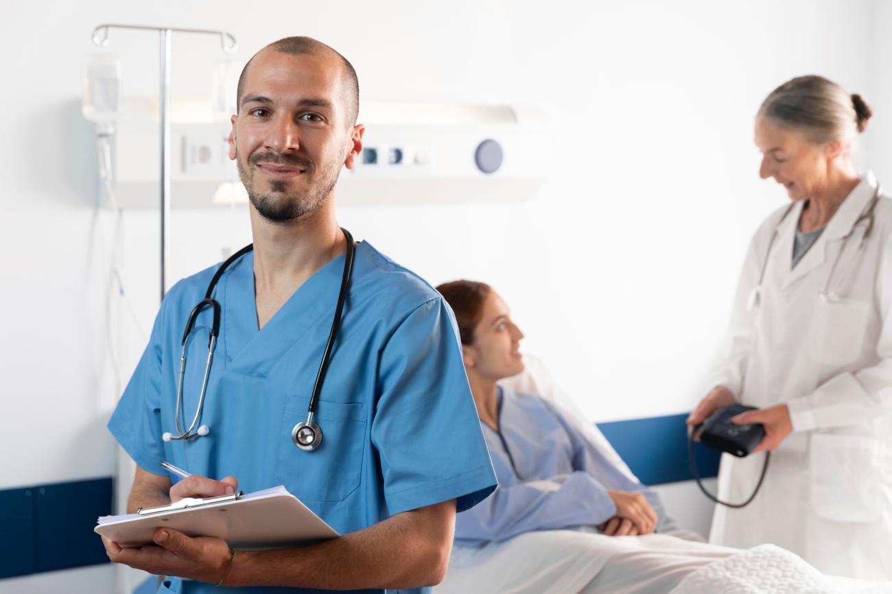 A male nurse smiles at the viewer while a doctor checks in with a patient in the background. Image by Freepik.