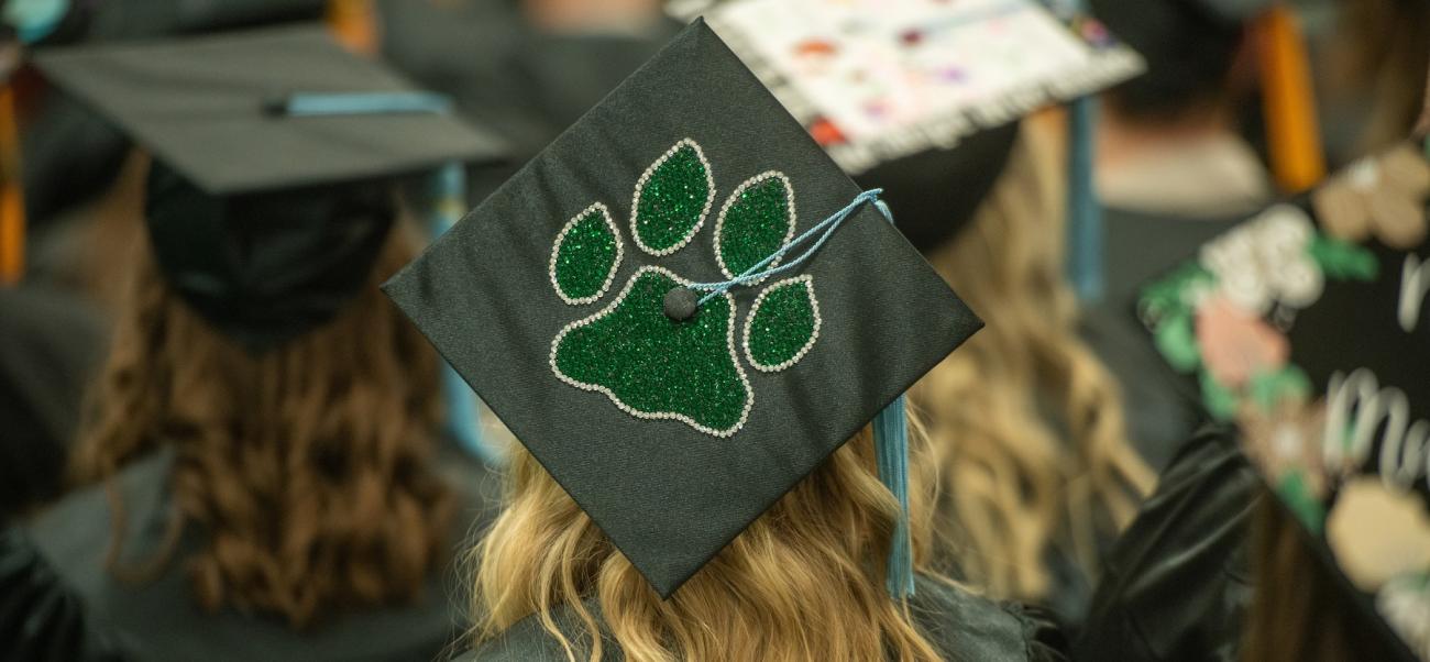 A graduate wearing a black mortarboard cap embellished with a green paw print