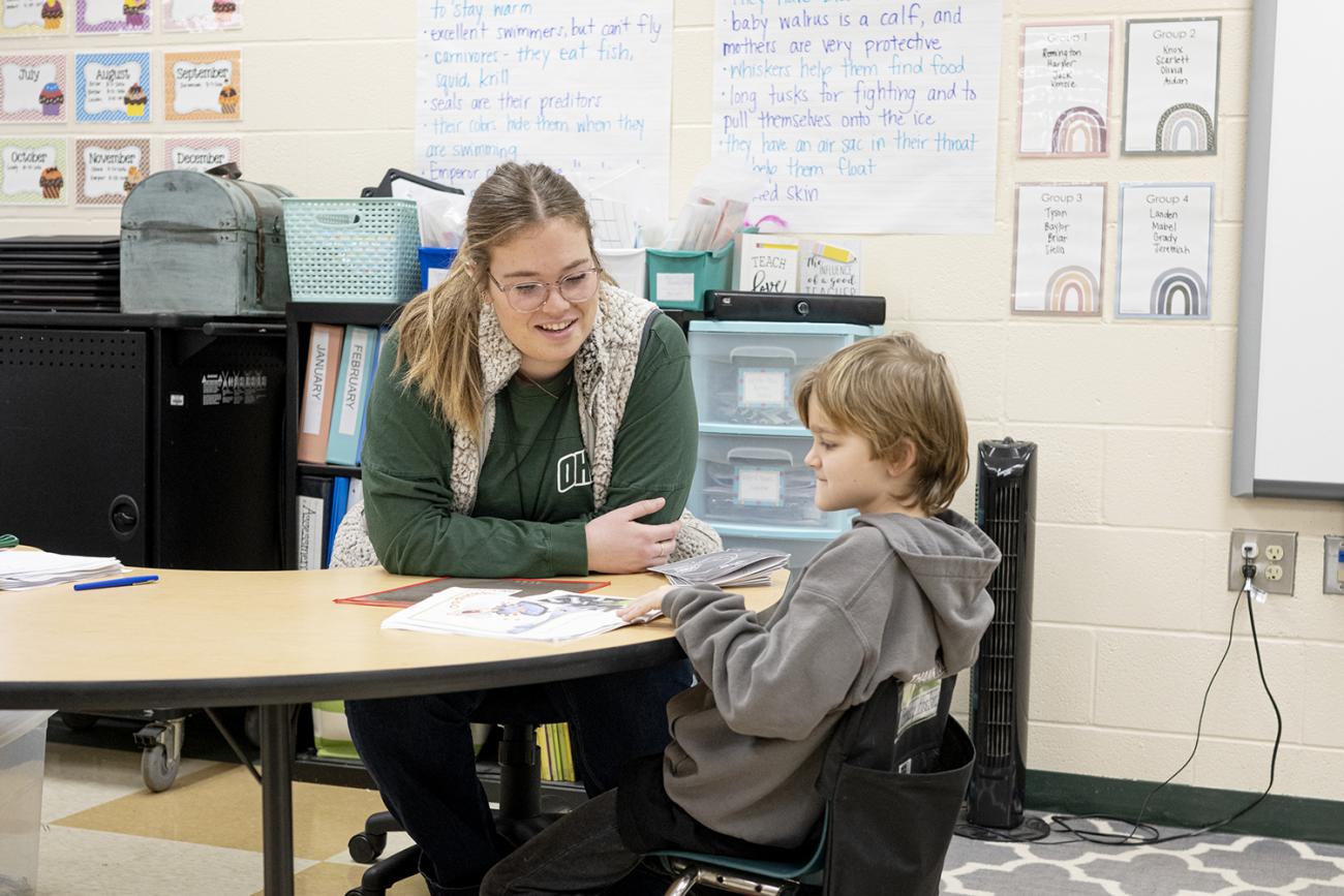 Woman sits with child listening to him reading in a class room