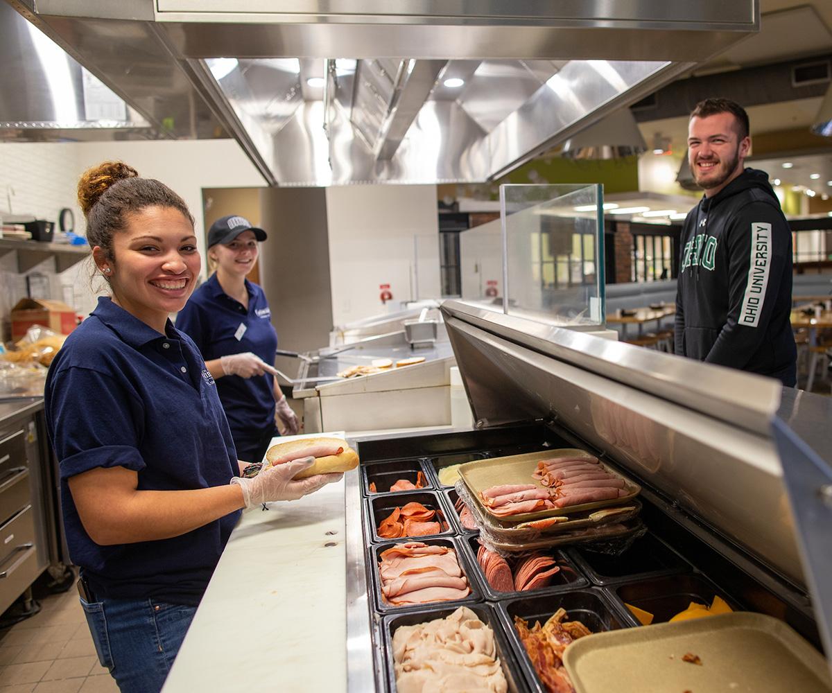 A student employee makes a sandwich in a dining hall for another student.