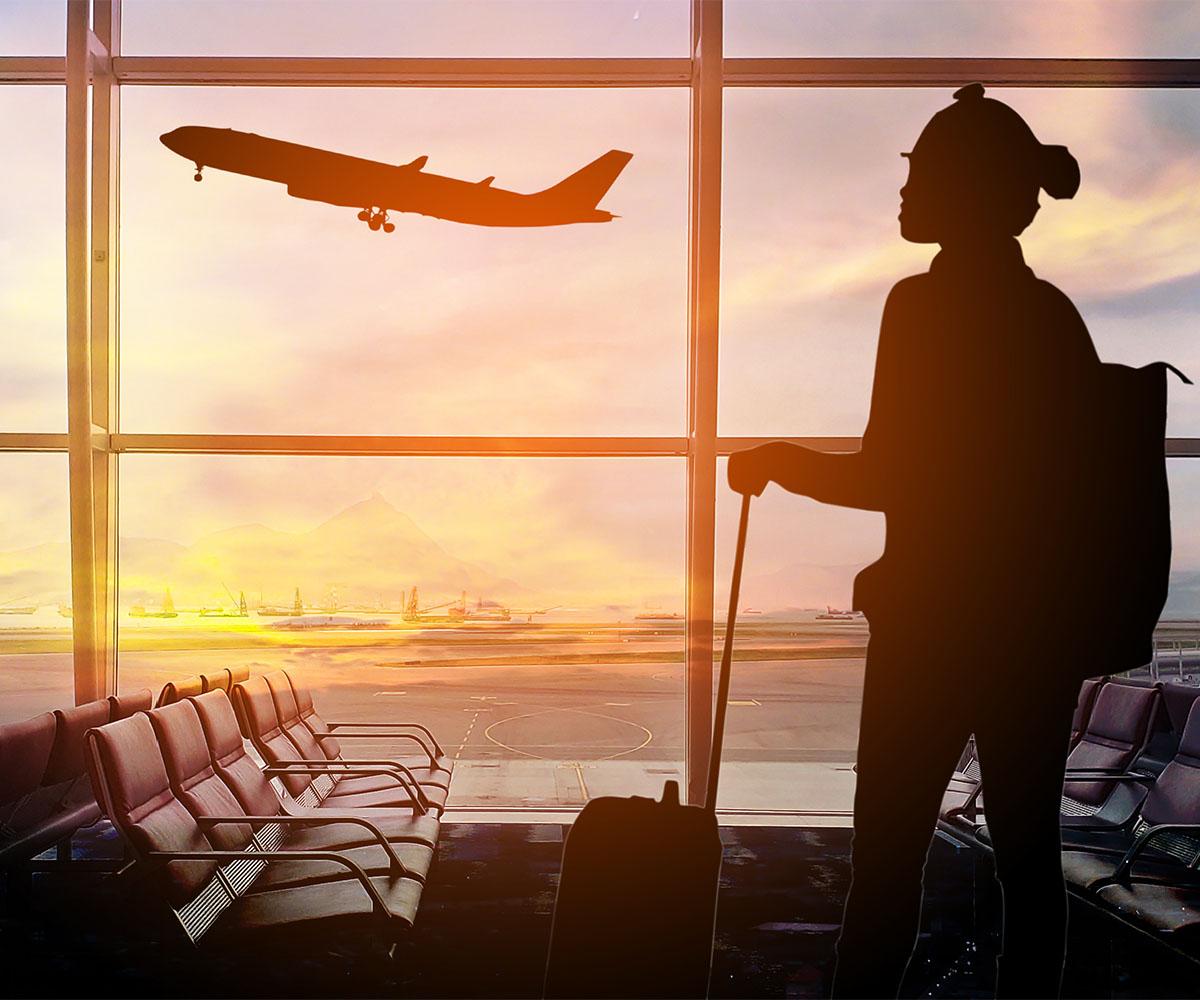 A person stands by a window in an airport watching a plane ascend above a sunrise.