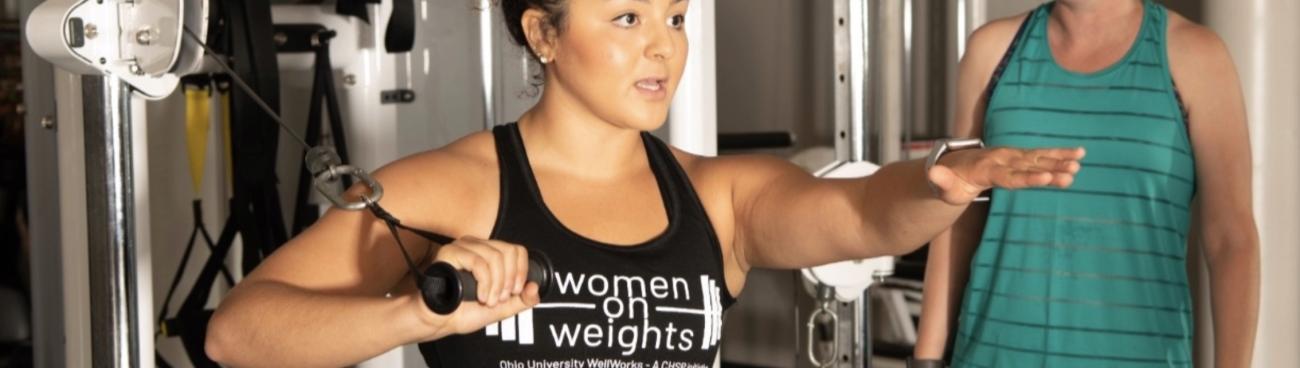 Woman using weight machine while wearing Women on Weights shirt