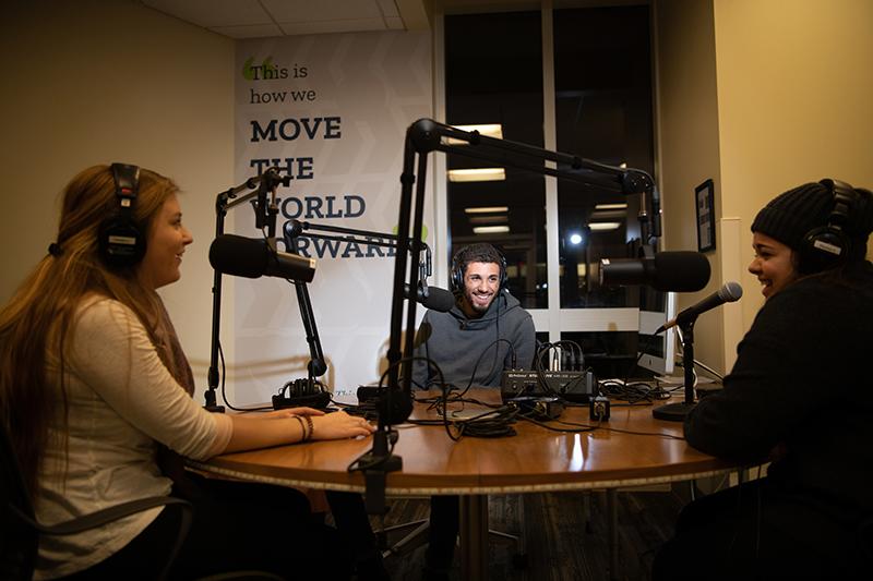 Three students sit in a podcast room, speaking into microphones and wearing headsets