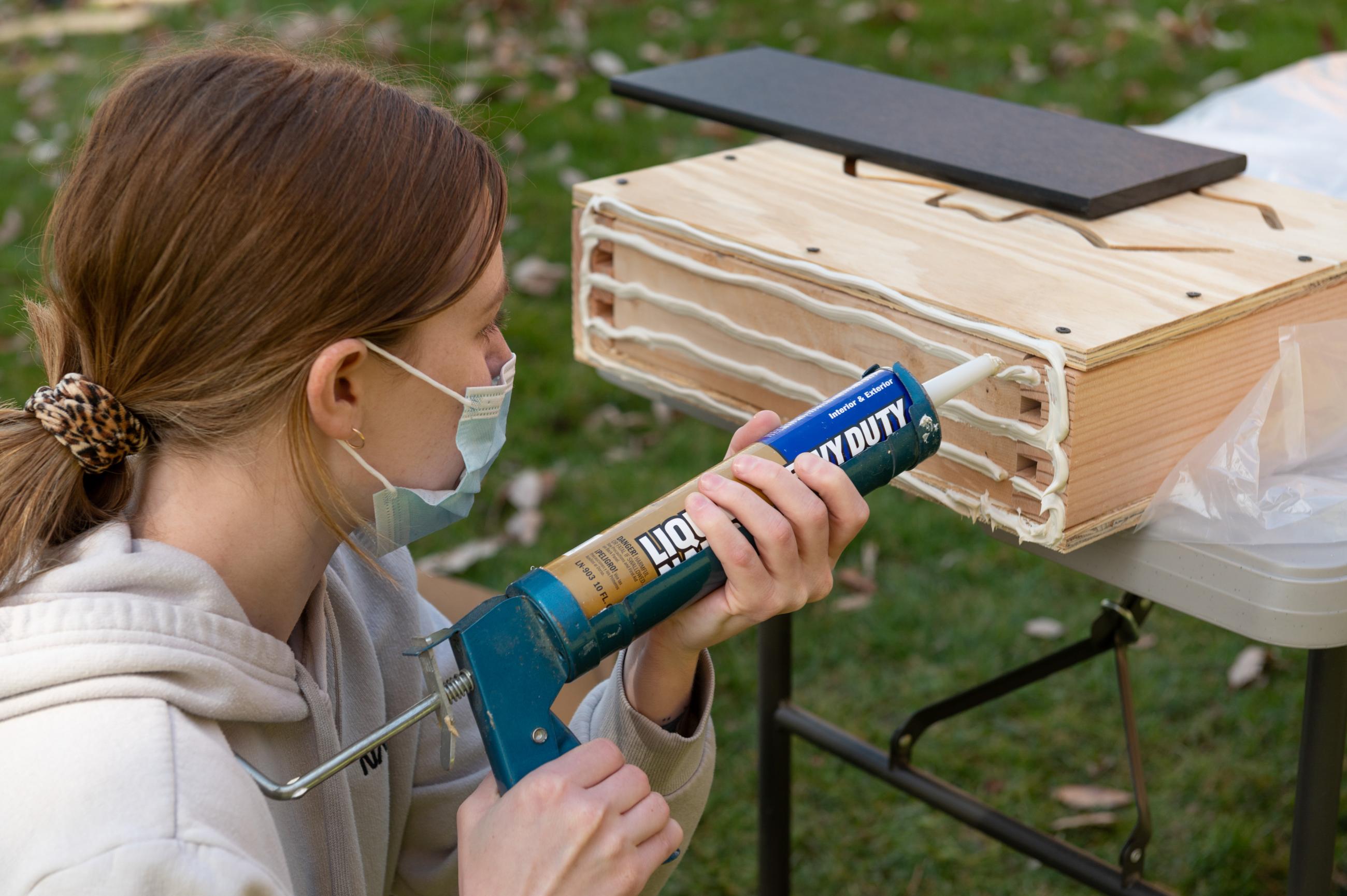 A woman uses liquid nails to attach the roof on a bat house.