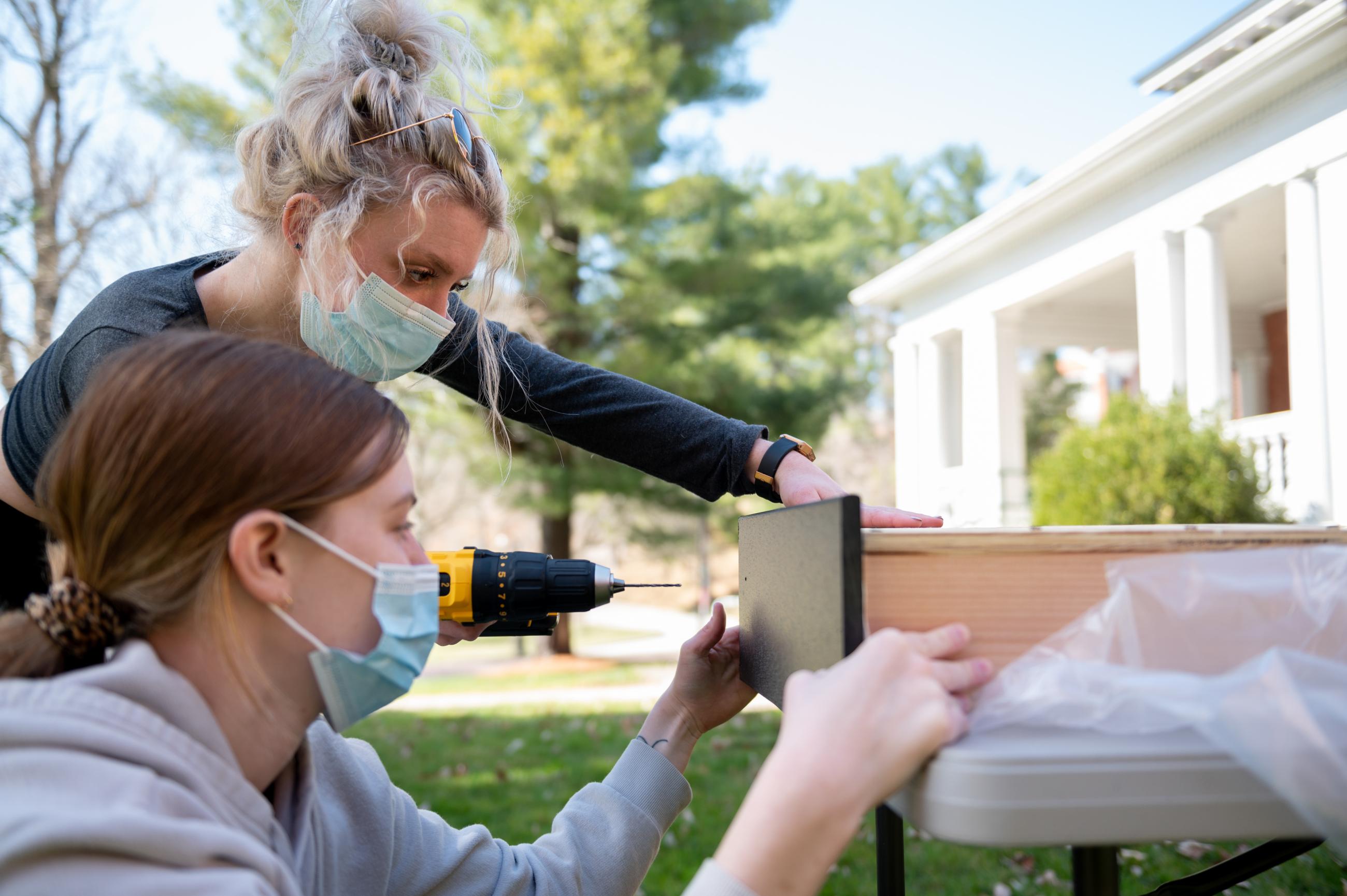Two woman use a power drill to attach the top piece to a bat house.