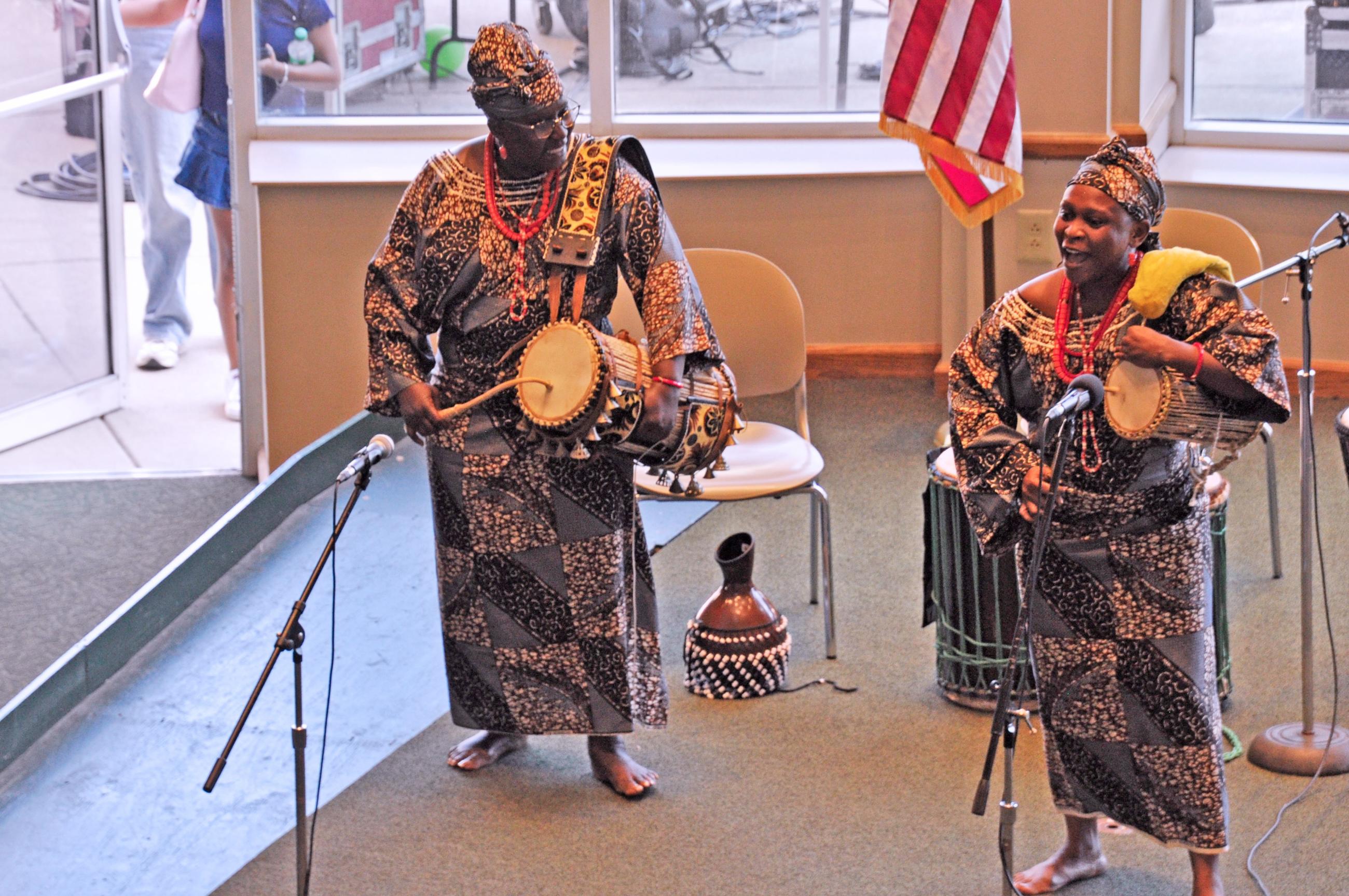 Two women in traditional African dress singing and playing drums