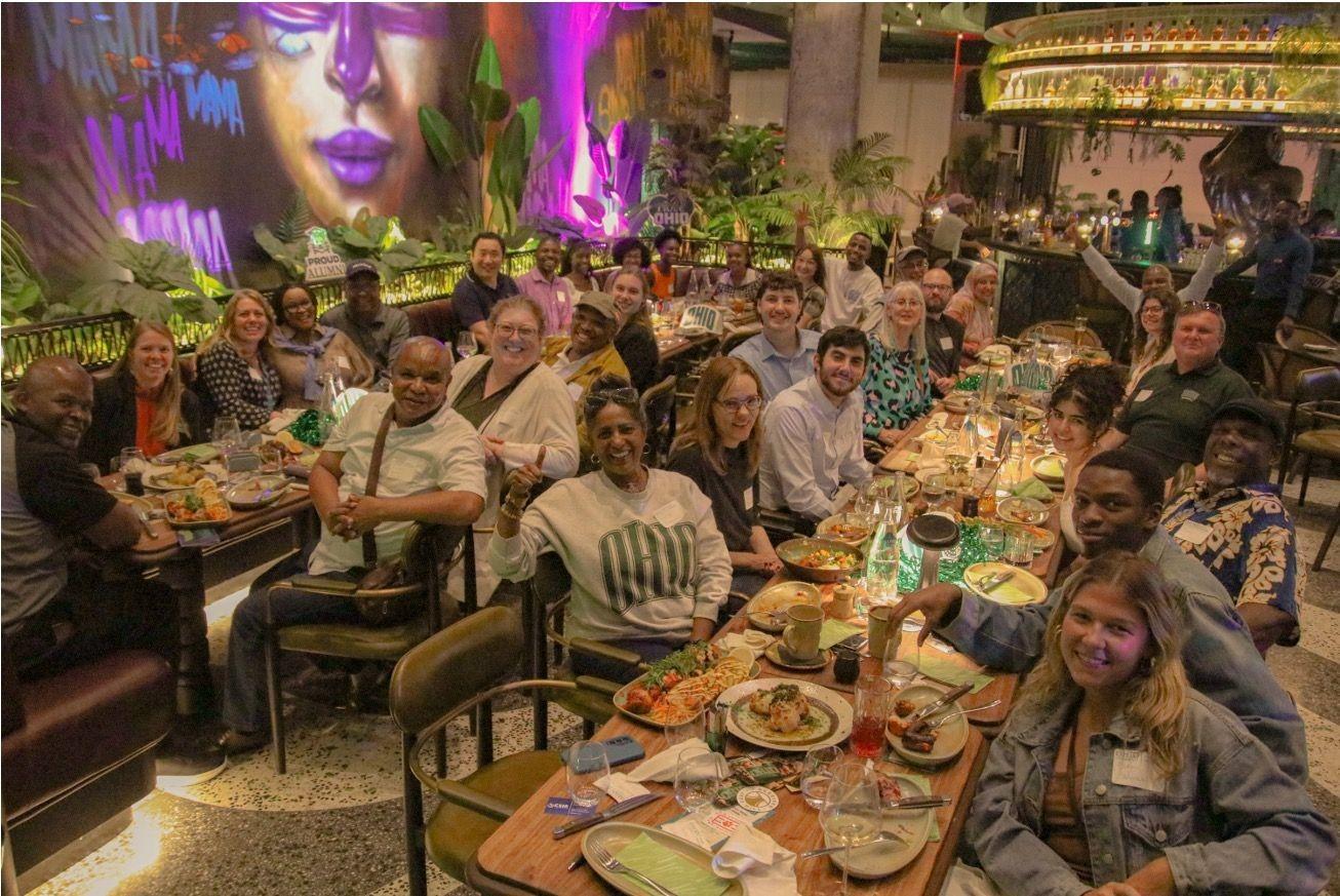 Large group of alumni sitting around long dinner tables eating meal in a restaurant.