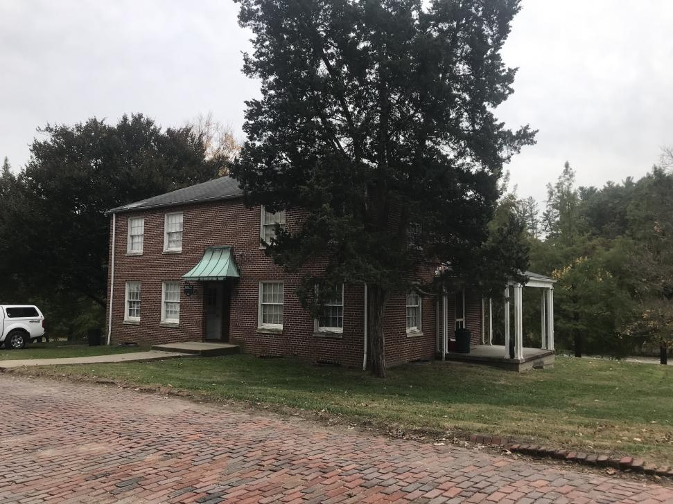 2-story rectangular brick building with front door looking out onto a brick street, with a side porch, and with a evergreen tree close to the corner of the building