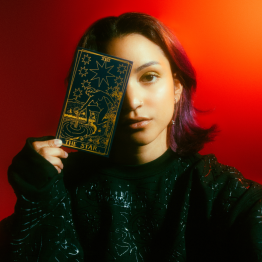 Headshot of Ambar Lucid covering her face with a tarot card with red background