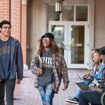 Two students walking and smiling on campus past two other students on a bench