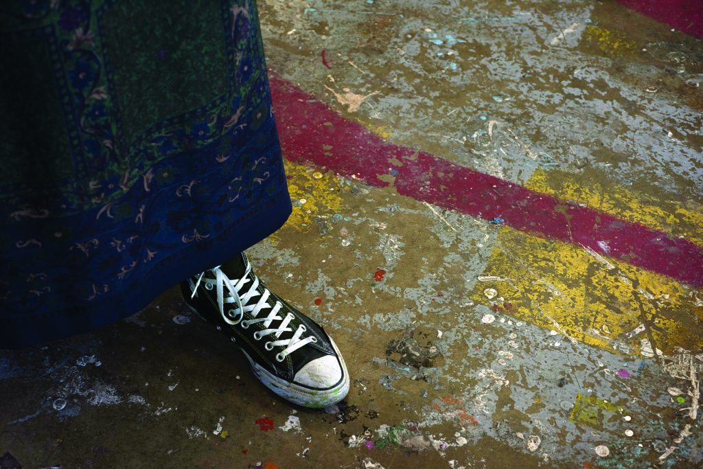 view of a person's feet, with painting smock visible, standing on a paint stained floor in a studio