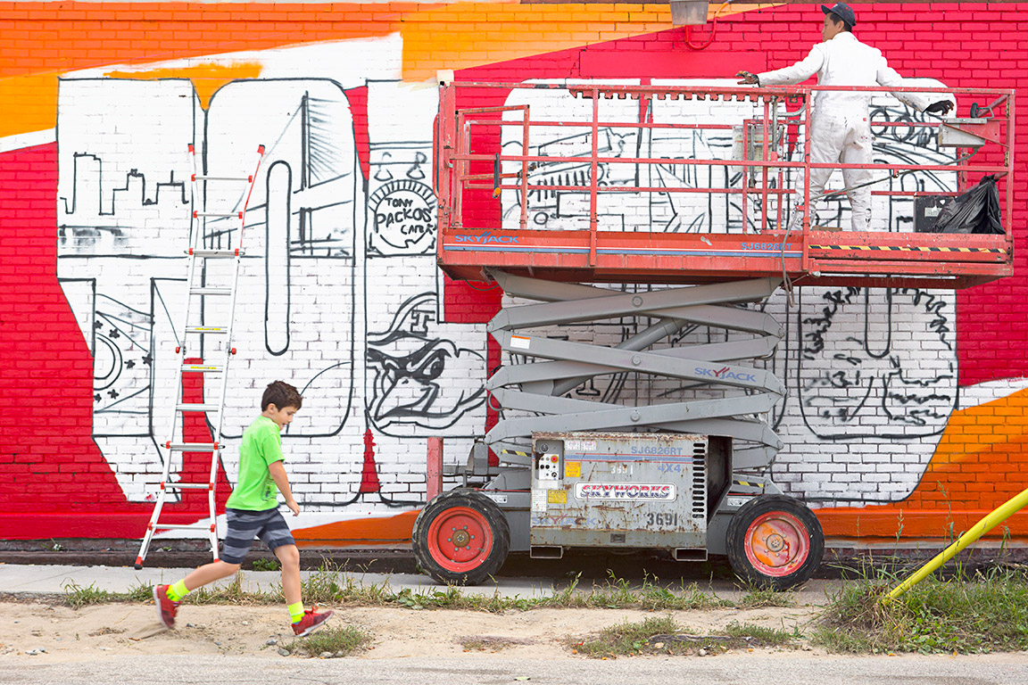 Child running in front of a building with a painted mural, with a painter standing on scaffolding in front of it